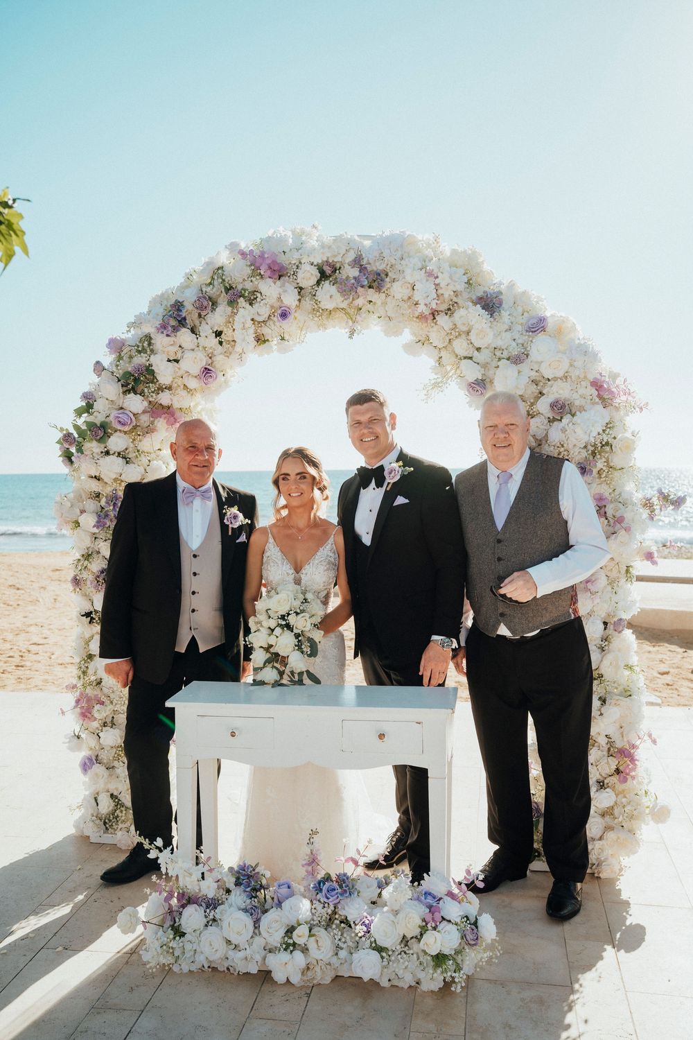 A wedding party gathers around an elegant white floral arch on a sunny beach for a signing ceremony.