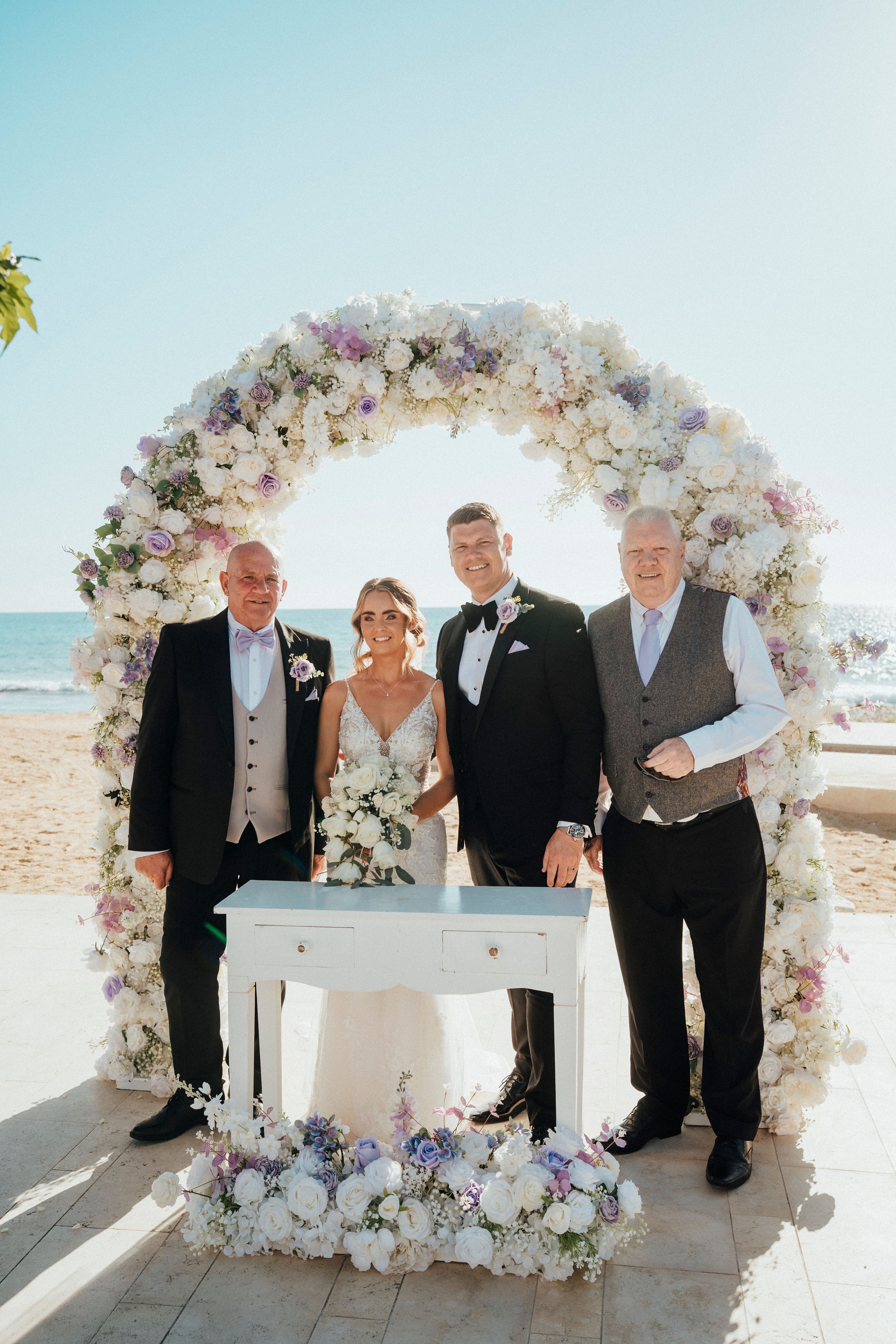 A wedding party gathers around an elegant white floral arch on a sunny beach for a signing ceremony.