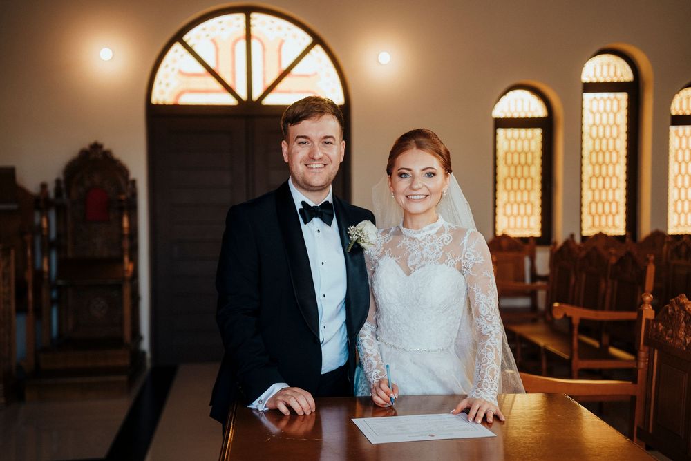 A couple signs marriage documents at a wooden table in a warmly lit church setting.