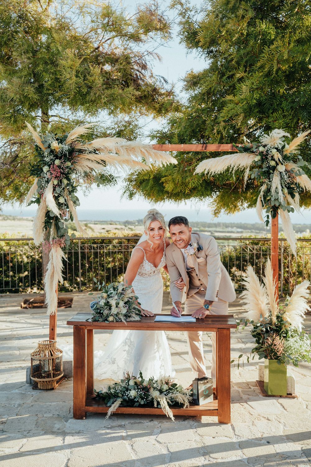 Rustic outdoor wedding ceremony setup with wooden benches and floral decorations under a tree in a sunny location.