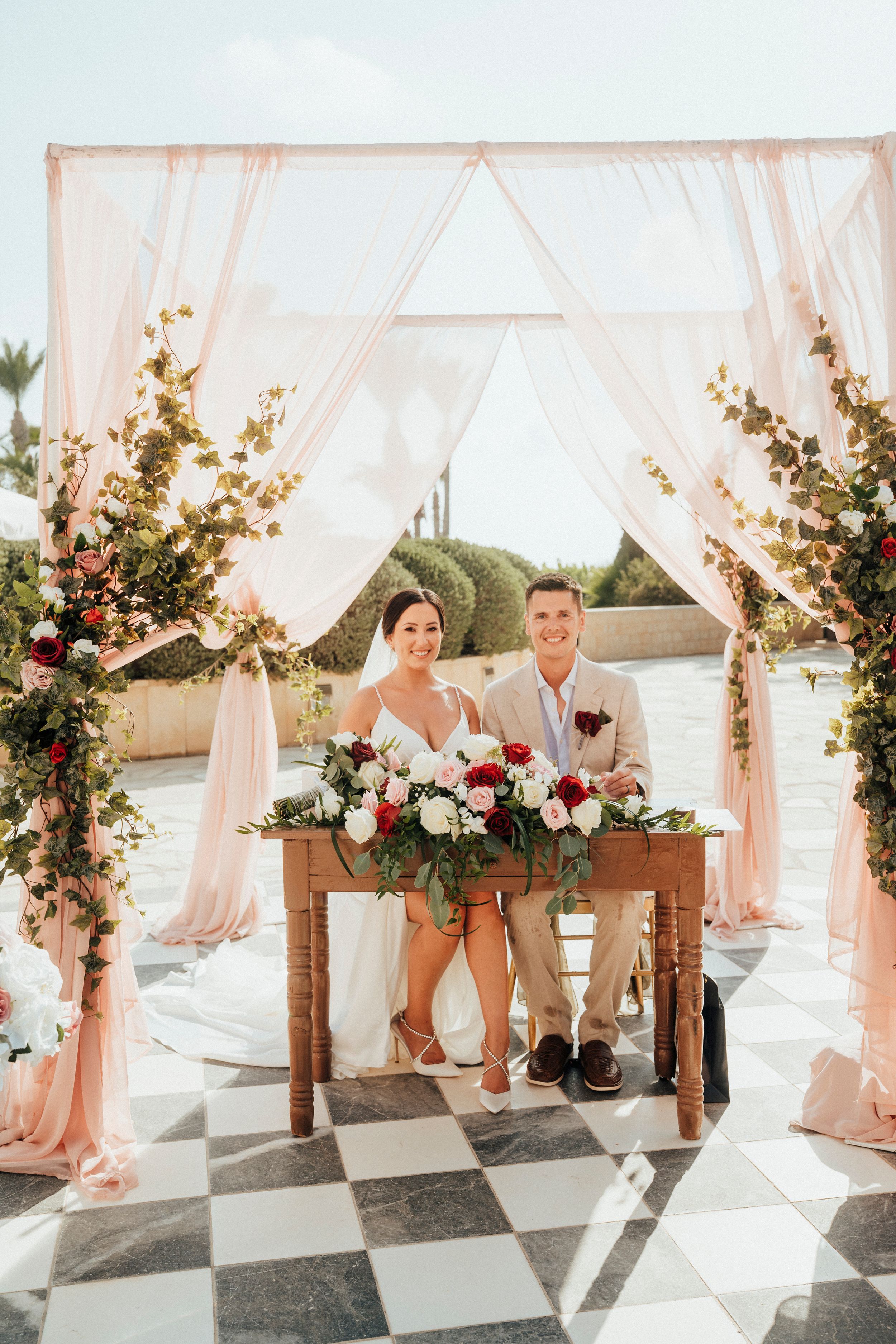 Wedding party poses in front of elegant white draped ceremony backdrop with pink and white floral arrangements.