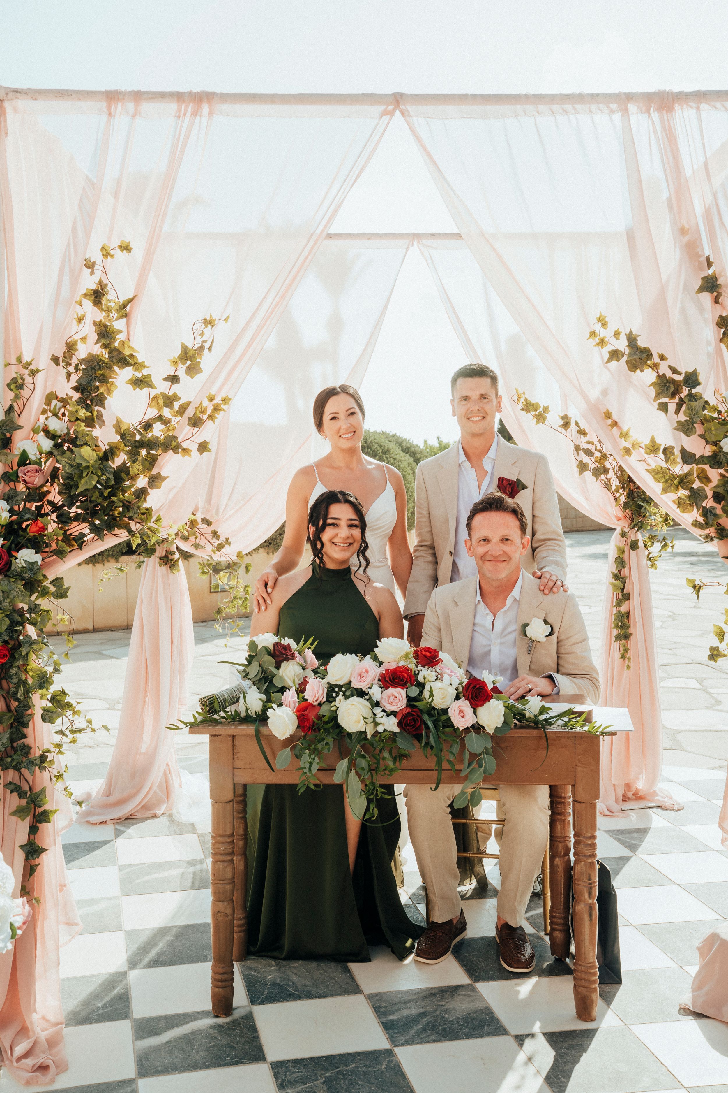 Wedding party gathered at rustic wooden table with floral arrangements under blush pink draped ceremony arch on checkered floor.