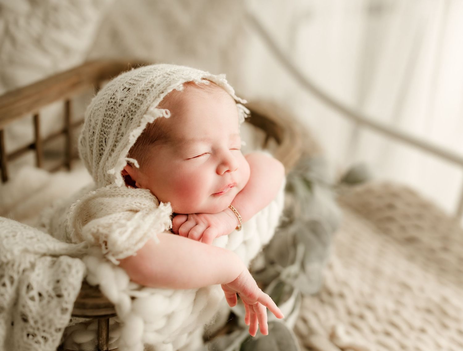 Peaceful sleeping newborn baby in cream knit bonnet posed in rustic wooden bowl with soft wraps.