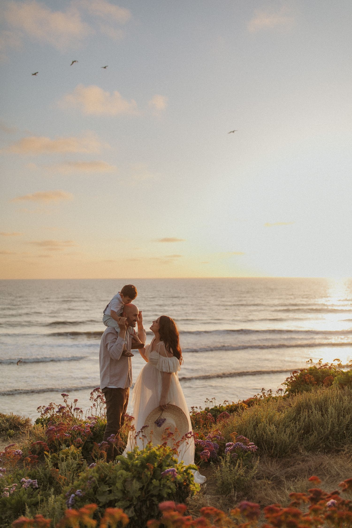 A couple stands on a coastal hillside surrounded by wildflowers during a golden sunset over the ocean. Carlsbad each maternity photography.