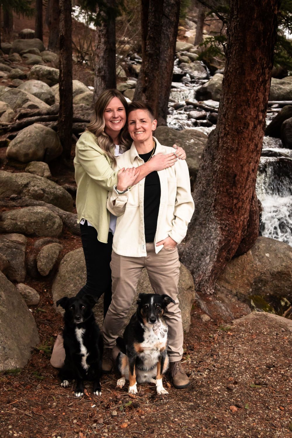 Couple poses with two dogs by a scenic forest waterfall during engagement photography in Idaho Springs, Colorado.