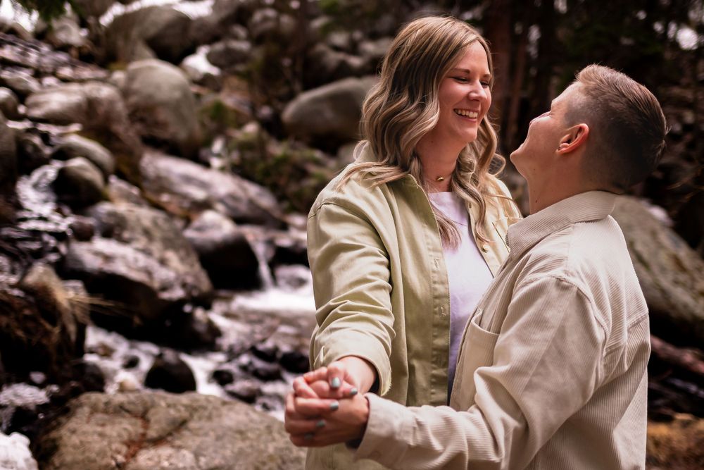 Couple sharing intimate moment by mountain stream in beige jackets during engagement photography in Idaho Springs, Colorado.