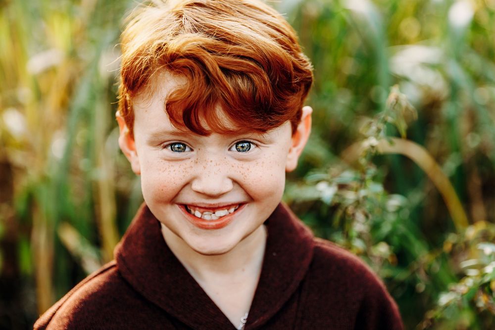 A child with bright red hair and blue eyes smiles while standing in a garden setting.