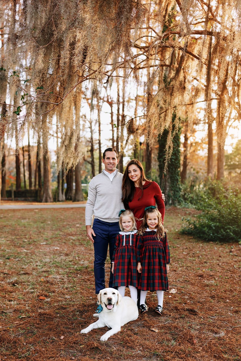 Family poses together with their white Labrador retriever under Spanish moss-draped trees at sunset.