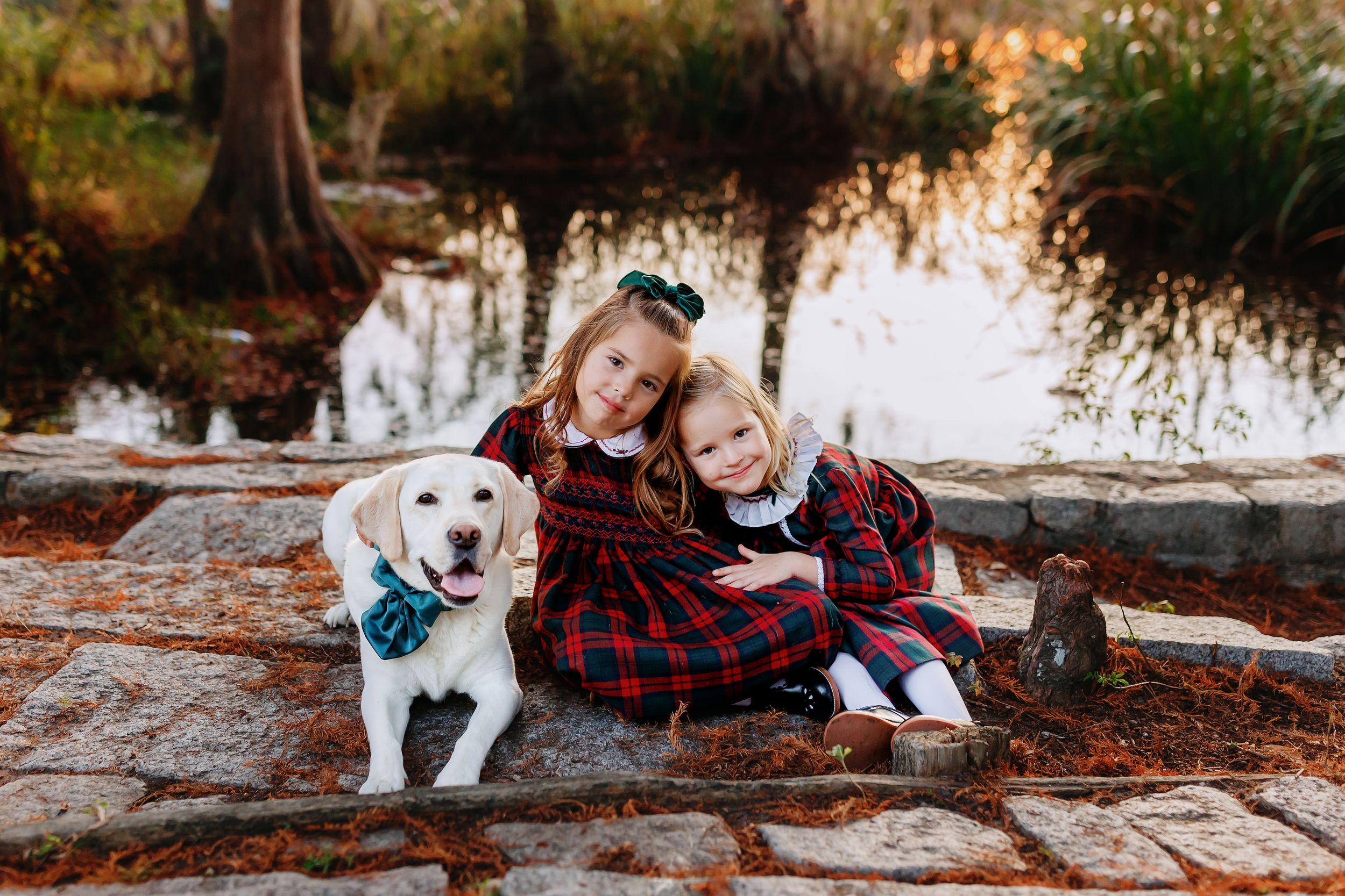 Two people in matching red plaid outfits sit on logs by a pond with their white dog wearing a blue bow tie.