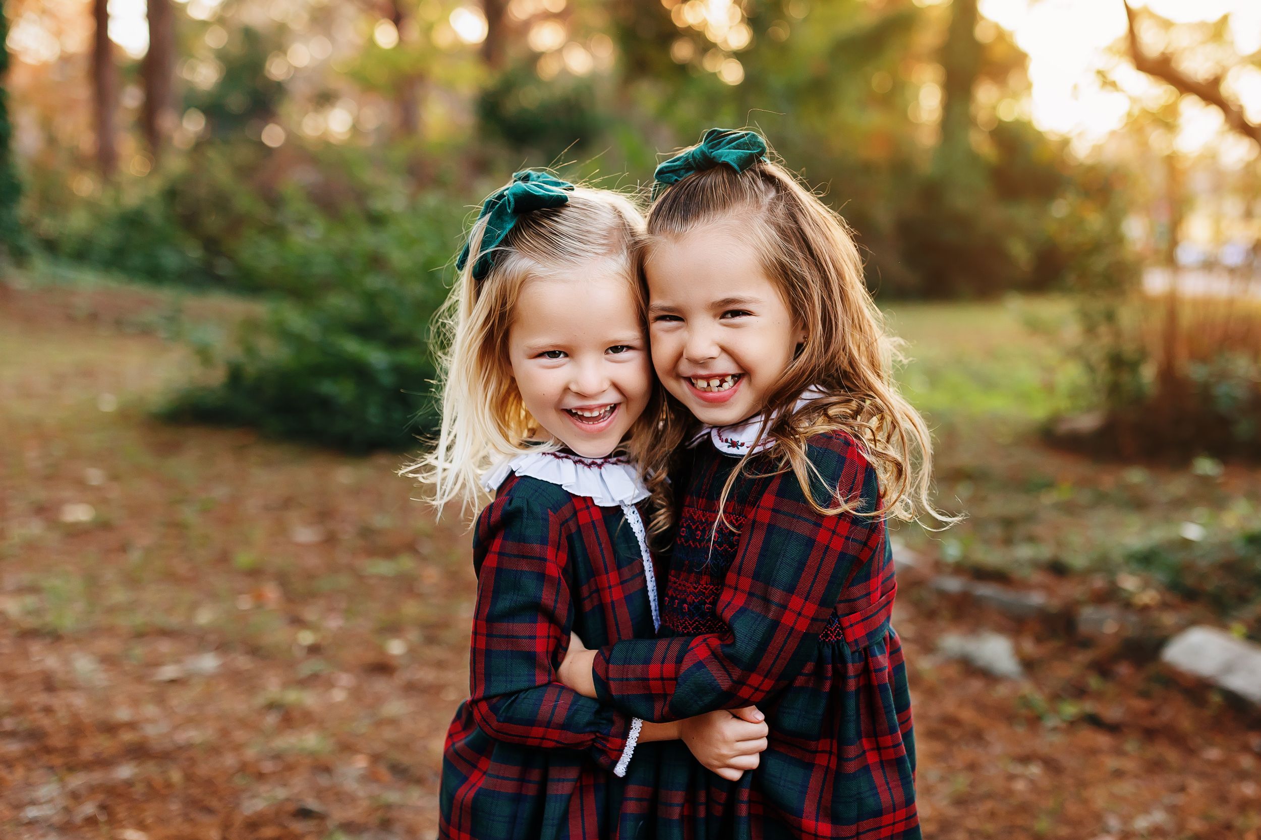 Two children wearing matching red plaid dresses and teal hair bows laugh together outdoors during golden hour.