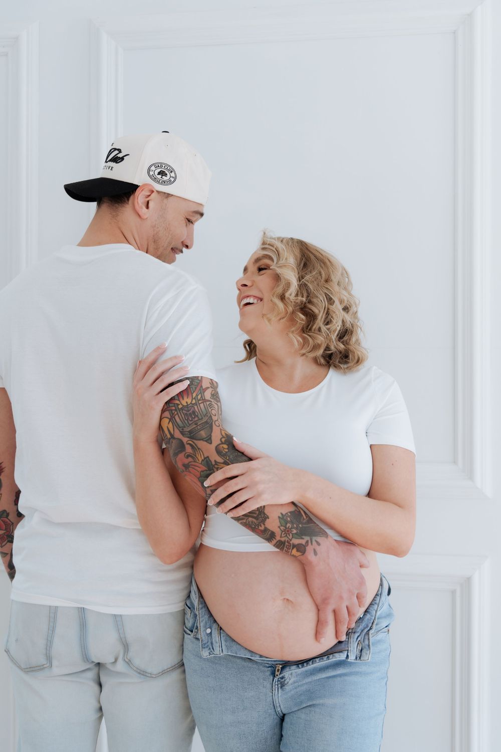 Tattooed pregnant couple in white t-shirts smiling at each other, woman cradling baby bump, against white wall.