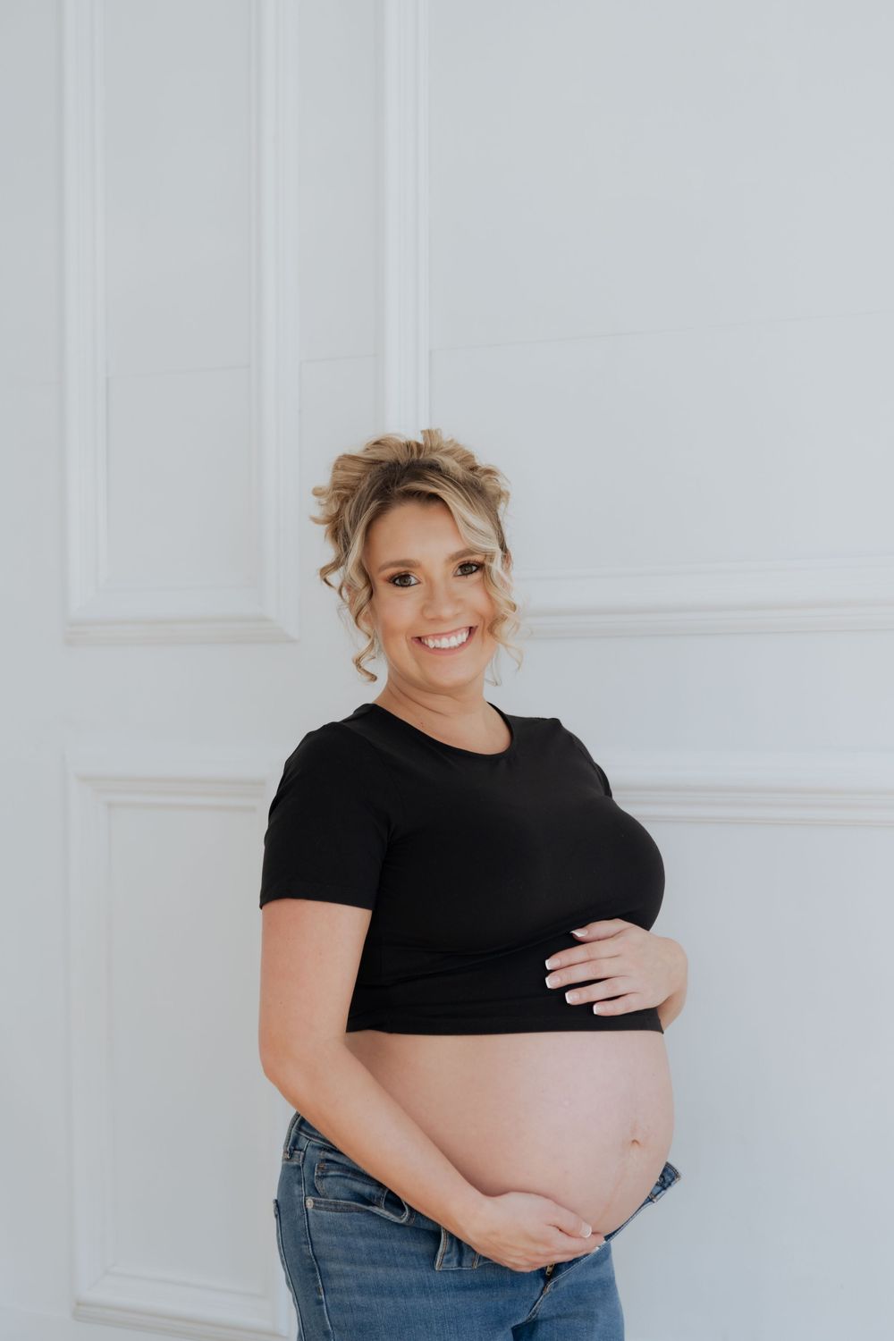 Smiling pregnant woman in black crop top and jeans cradling baby bump against white wall.