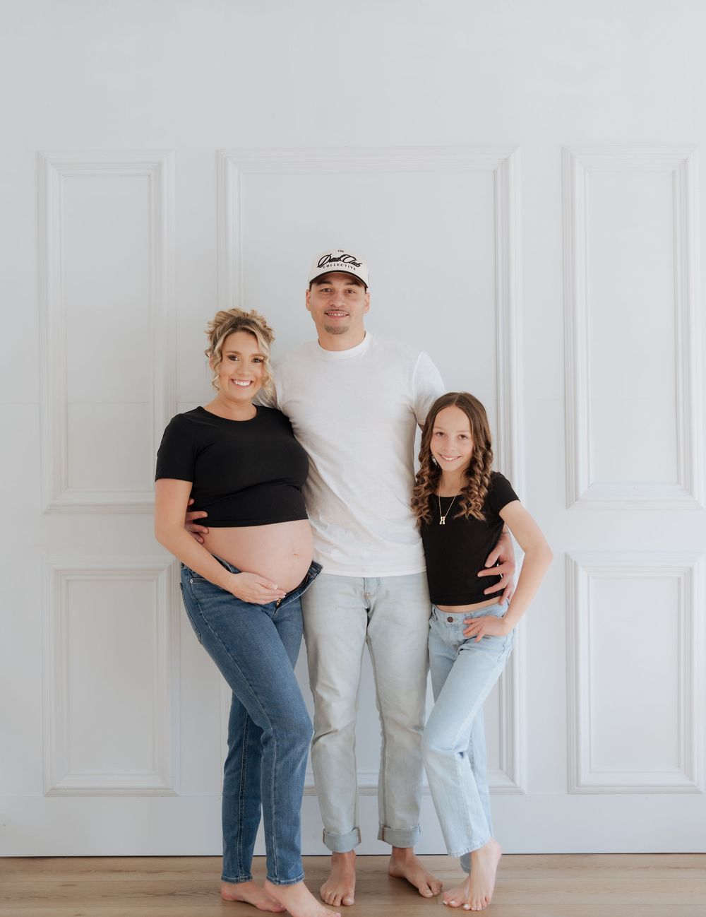 Pregnant woman poses with man in white cap and young girl against white paneled wall in maternity photo.