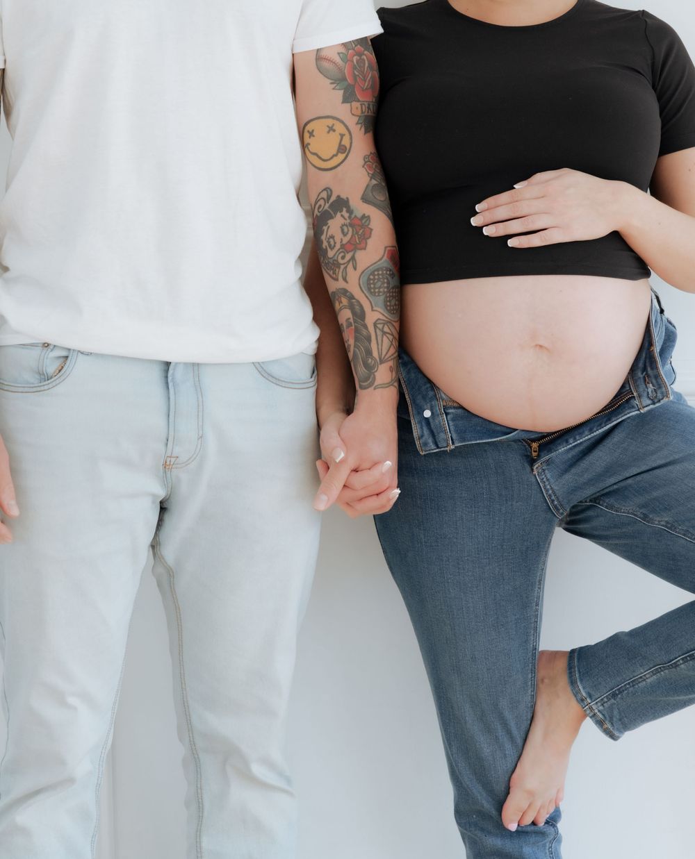 Pregnant woman in black crop top and jeans holds hands with tattooed partner against white wall.