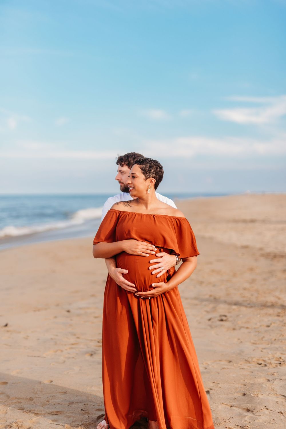 Pregnant couple embracing on sandy beach, woman in rust off-shoulder maxi dress, ocean waves and blue sky behind them.