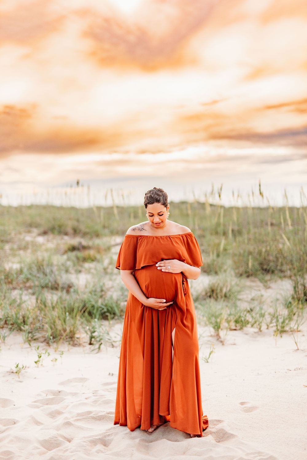 Pregnant woman in rust orange off-shoulder maxi dress cradling baby bump on sandy beach at golden sunset.