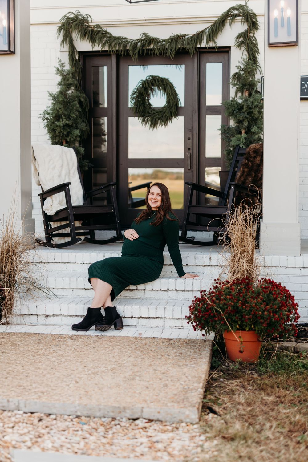 Person in dark clothing poses artistically on white stone steps of a building with evergreen decor.