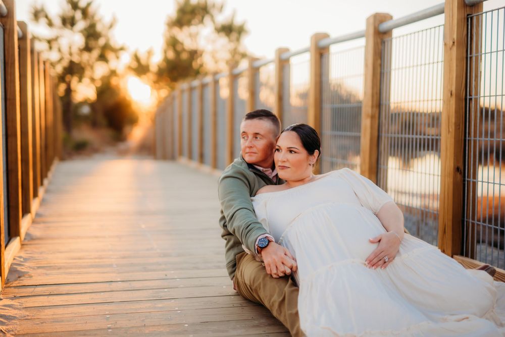 A couple sits together on wooden boardwalk during sunset with warm lighting and railing behind them.