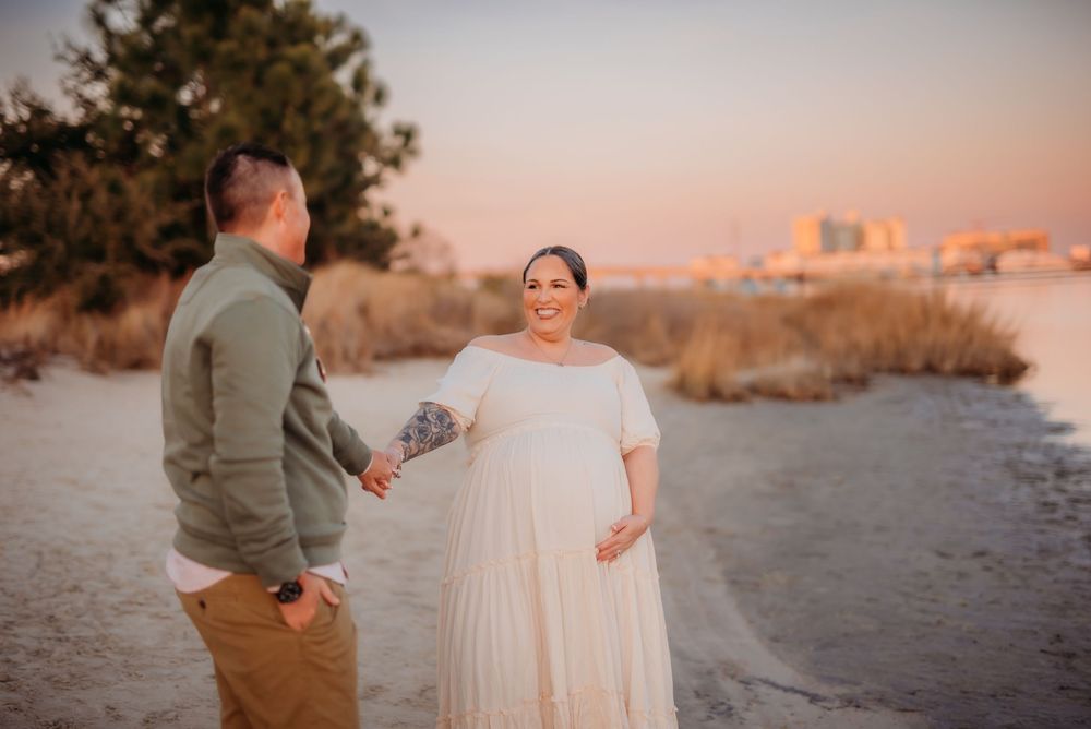 Pregnant woman in white gown holds flowers with partner on sandy beach at sunset.