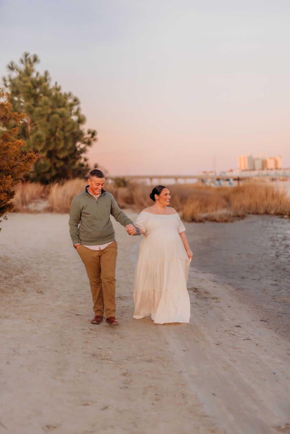 Couple walks hand-in-hand on sandy beach path at sunset, woman in flowing white gown, man in green sweater.