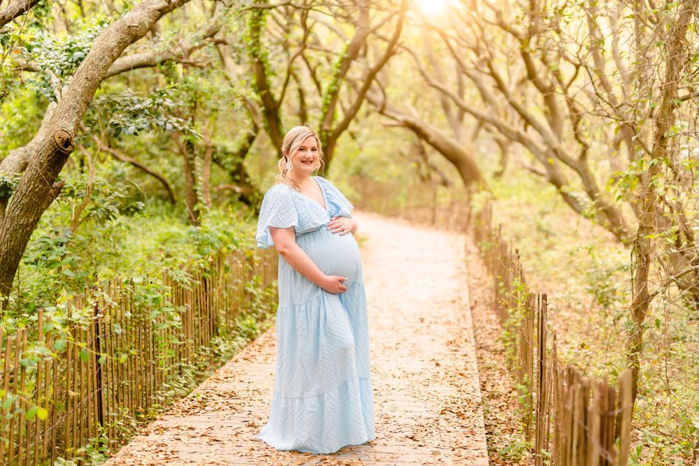 Expecting mother in flowing blue dress poses on sunlit forest path.
