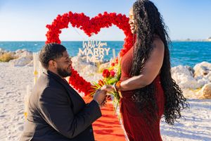 Man proposing to woman on beach with red heart floral arch and Will You Marry Me sign in background.