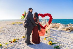 Couple embracing before a red rose heart arch on a sandy beach with sunflowers and ocean backdrop.
