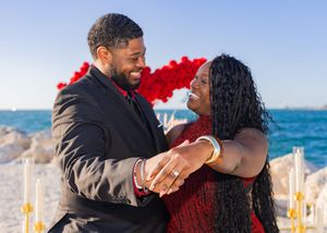 Couple sharing a joyful engagement moment on a beach, showing off ring, with red rose heart backdrop.