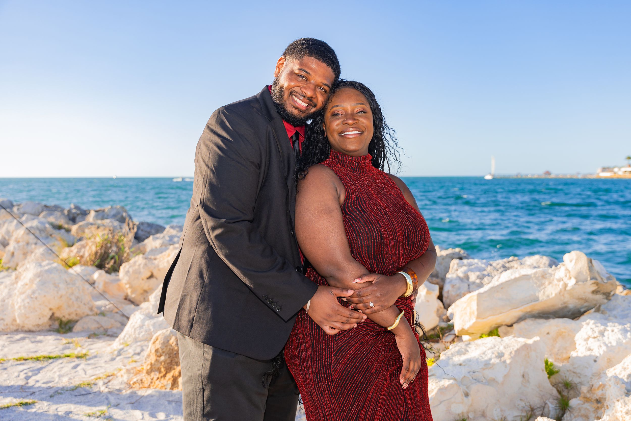 Couple in formal attire embracing and smiling on rocky beach with turquoise ocean in background on sunny day.