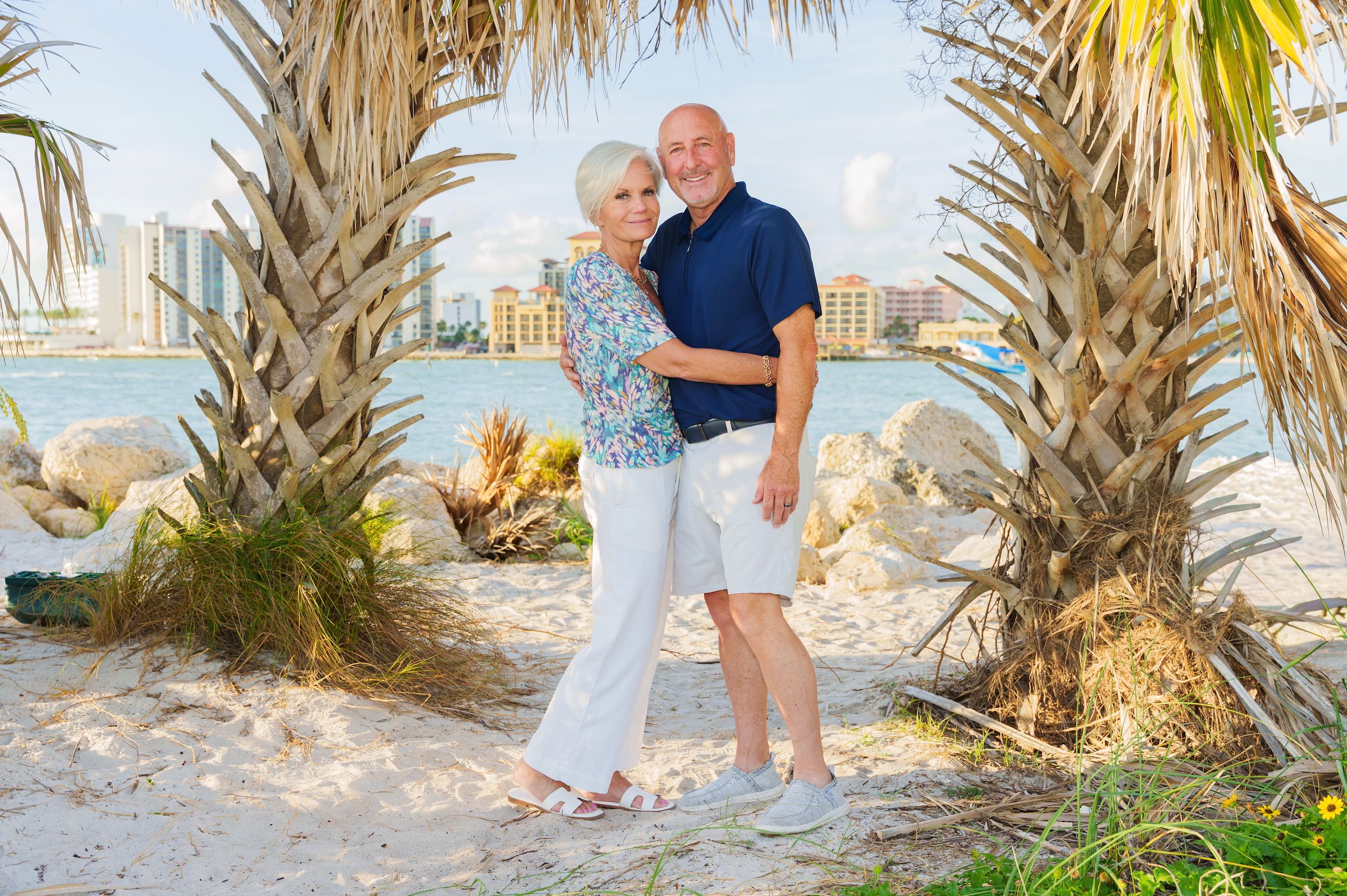 Couple posing together on a sandy beach with palm trees and city skyline in the background.