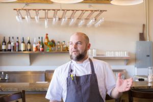 Restaurant staff member in white chef coat and gray apron stands in professional kitchen.