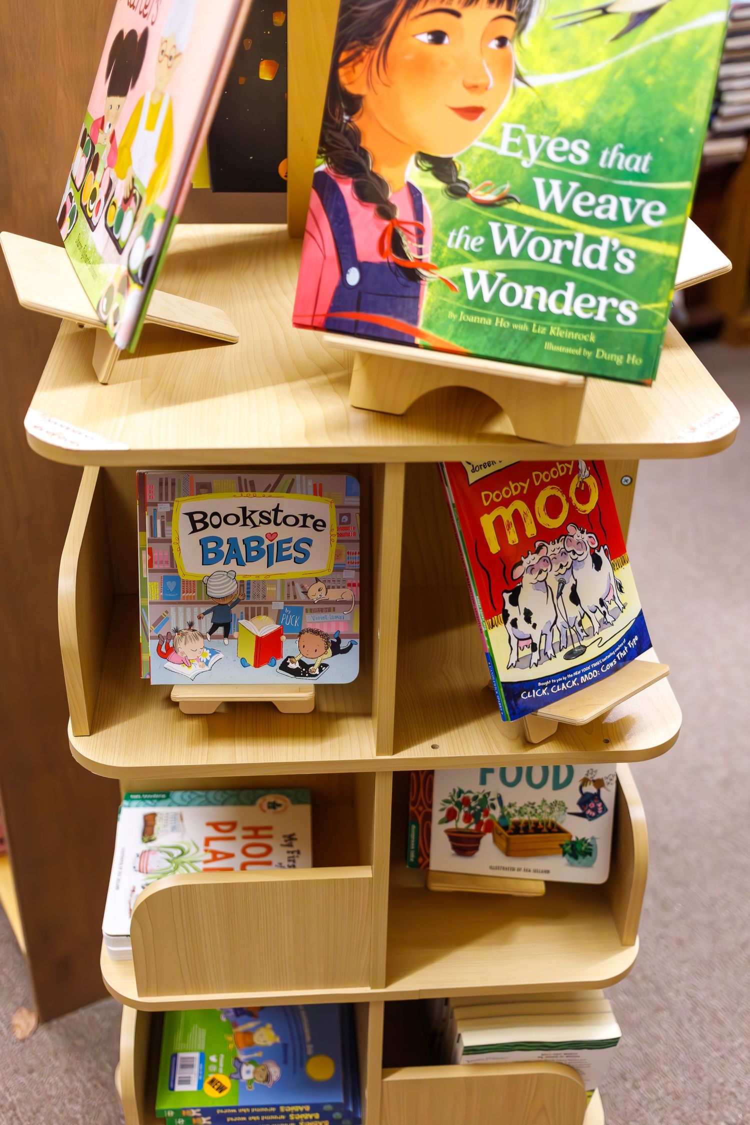 Children's books displayed on a wooden shelf including 'Eyes that Weave the World's Wonders' and other titles.