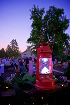 A red camping lantern glows at dusk in a garden setting with trees silhouetted against a purple twilight sky.
