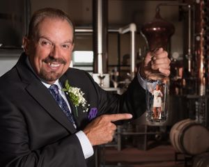 A suited businessman points to a bottled spirit inside a distillery with wooden barrels in the background.