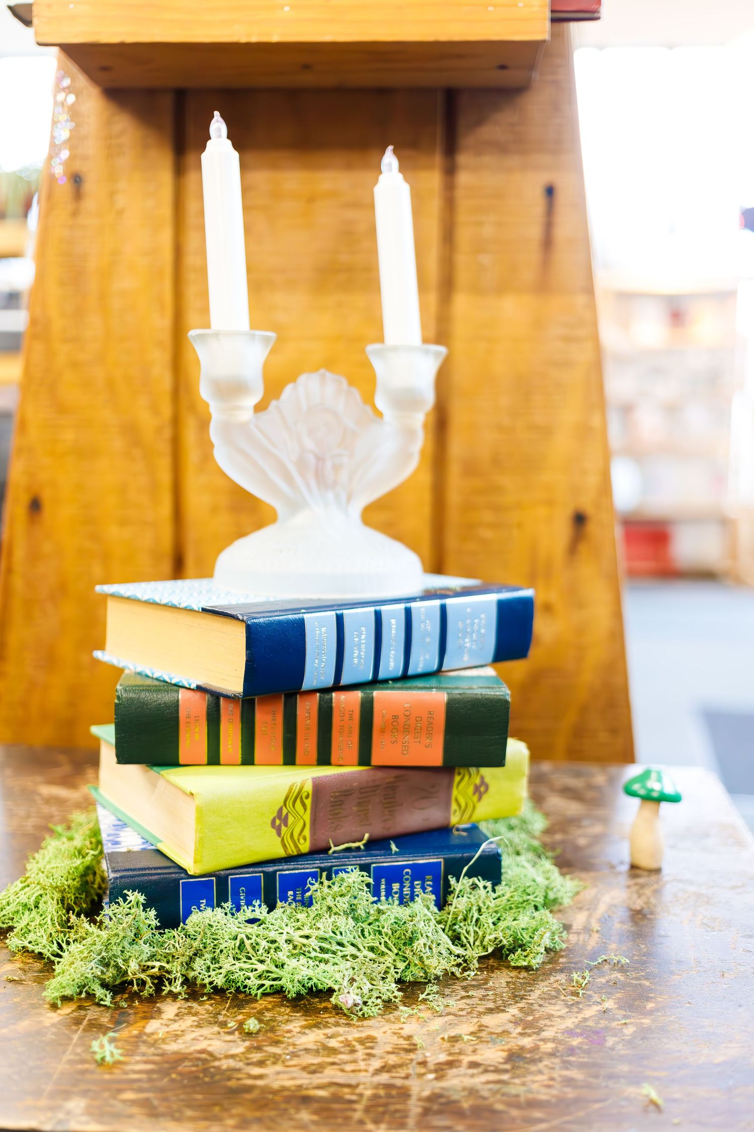 Stack of vintage books with white candle holder and moss decoration on wooden surface.
