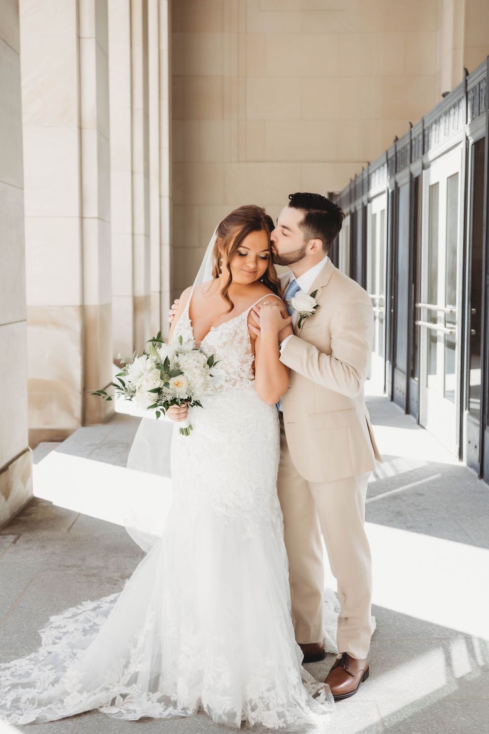 Intimate moment between newlyweds in formal attire against a light-colored building exterior.