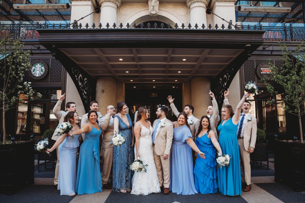 Wedding party in shades of blue and beige poses together under an ornate building entrance.