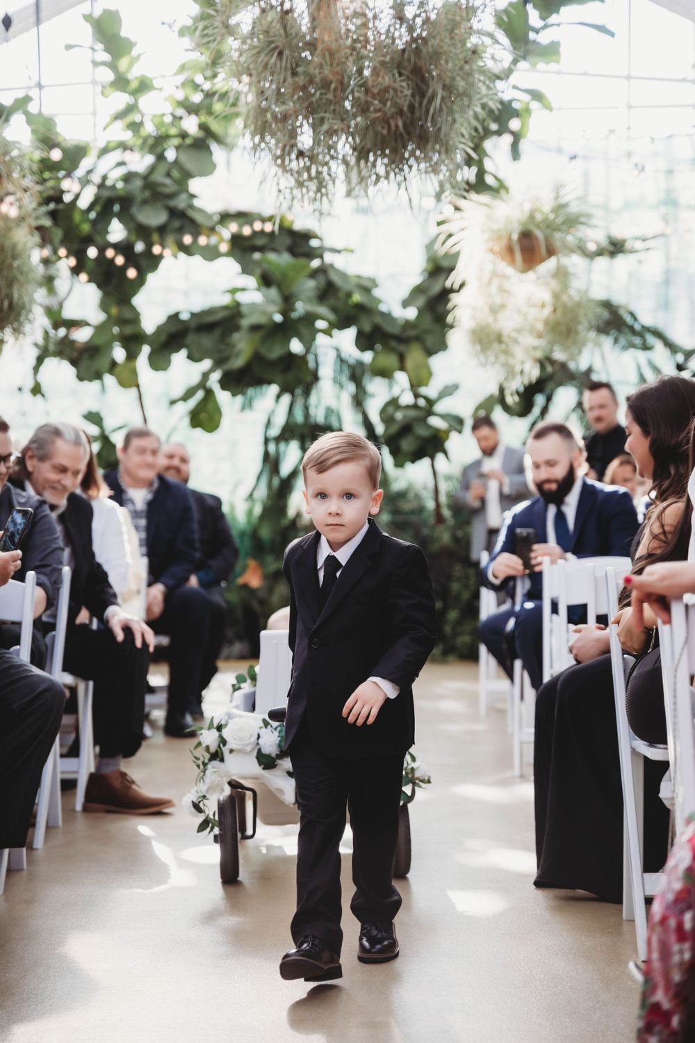 Young ring bearer in black suit walks down greenhouse ceremony aisle.