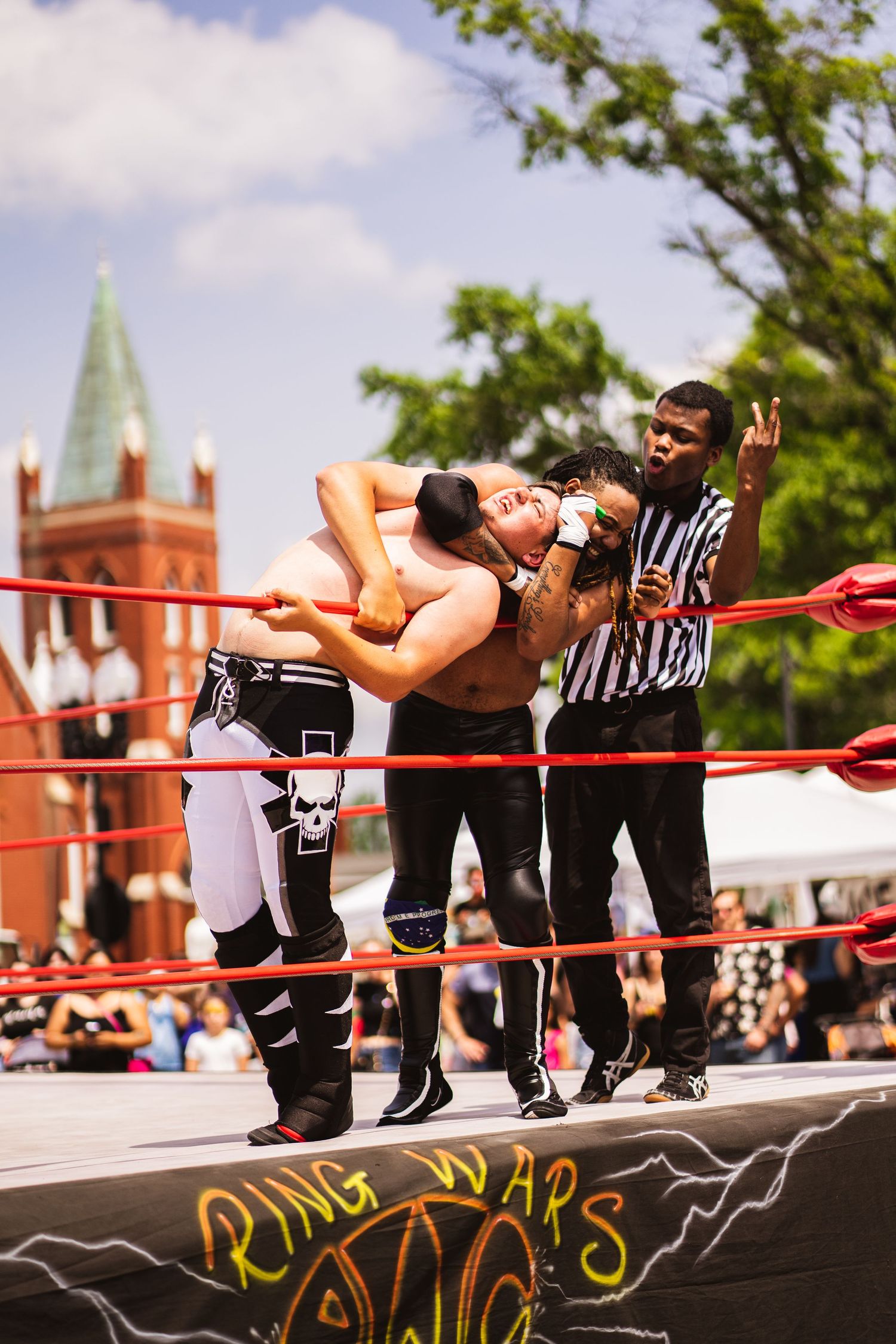 Professional wrestlers performing a high-flying move in an outdoor ring with church spires in background.