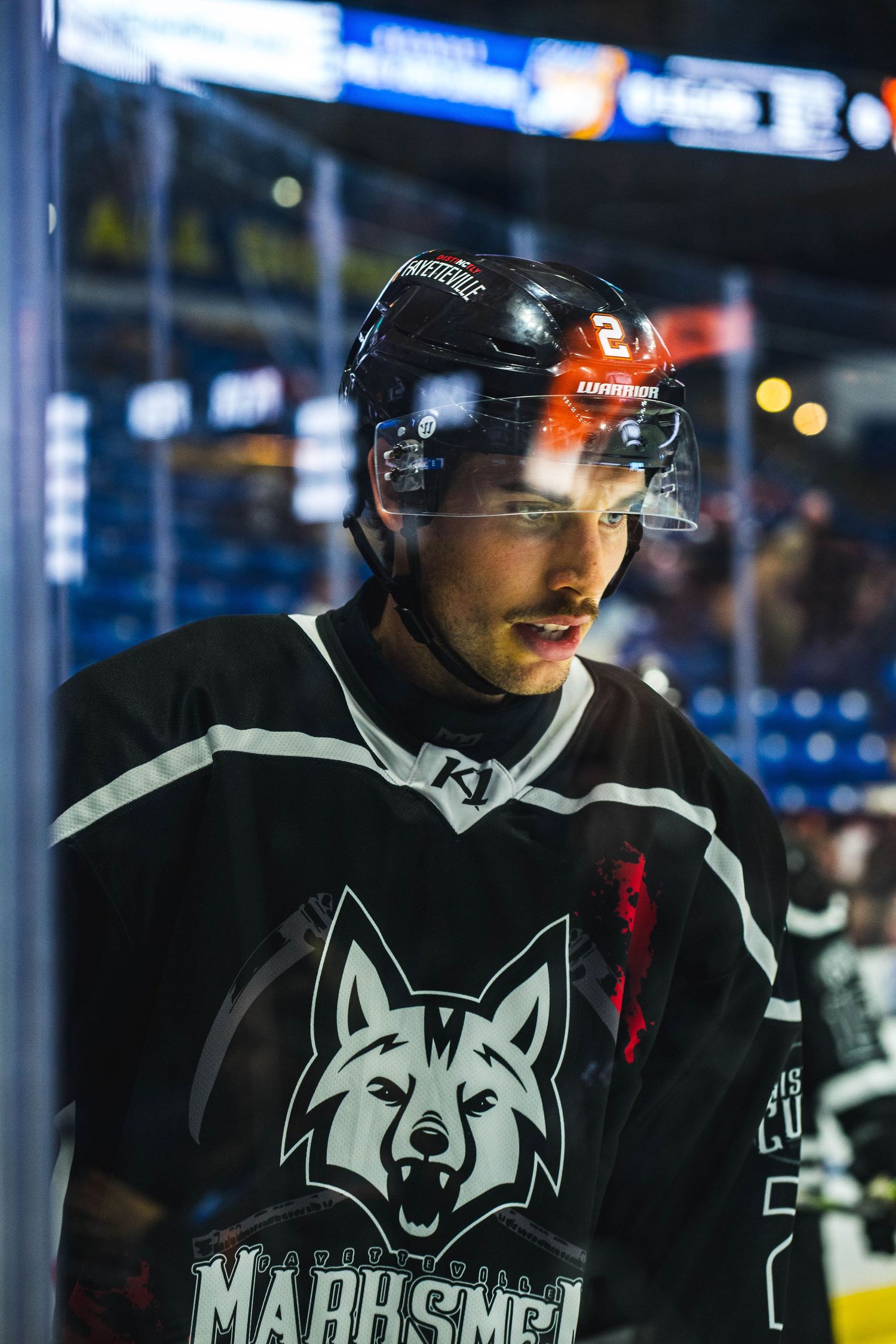 Hockey player in black Marksmen jersey standing near arena boards during game.