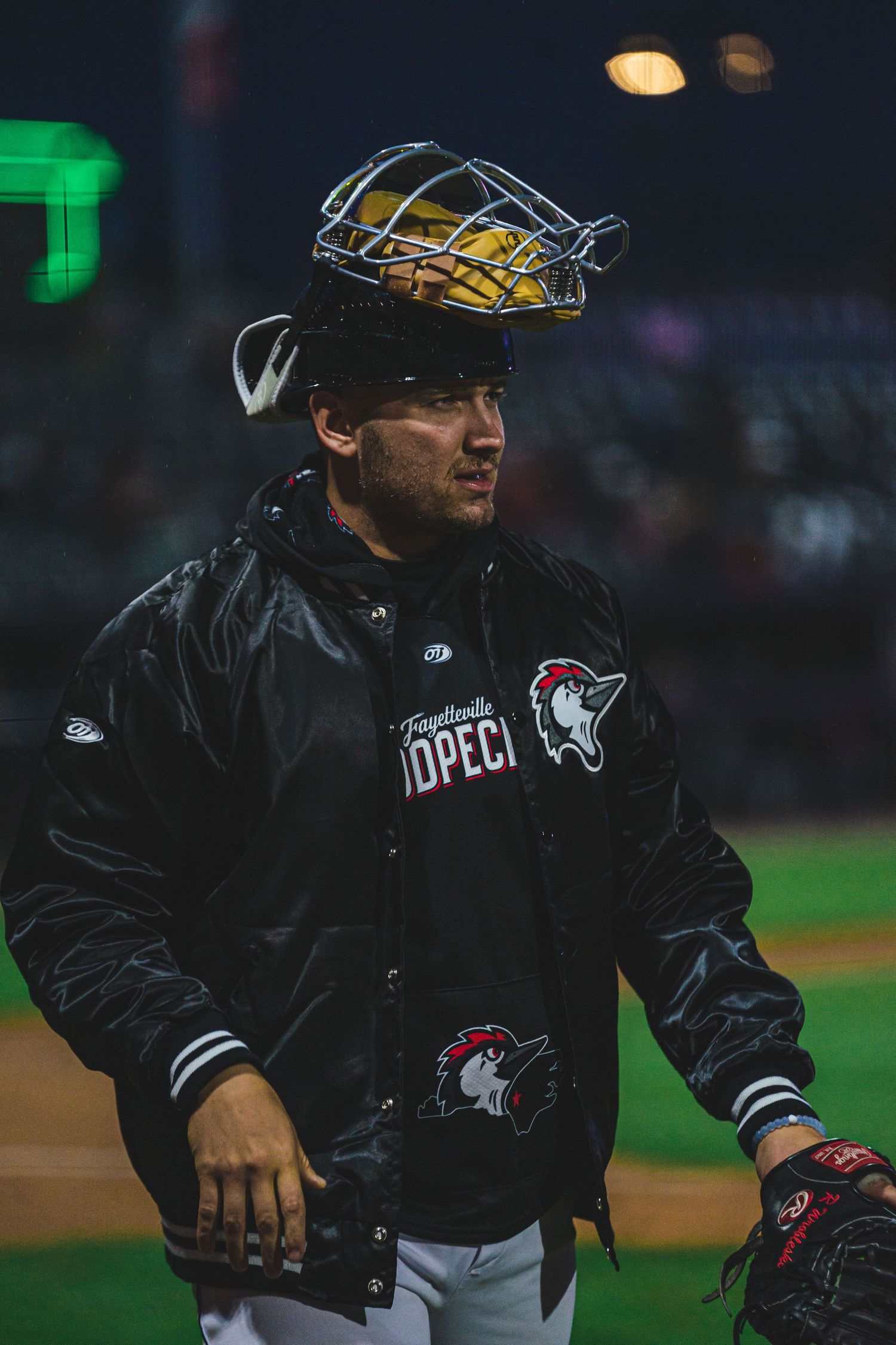 Baseball player in black team jacket with protective gear balanced on cap.