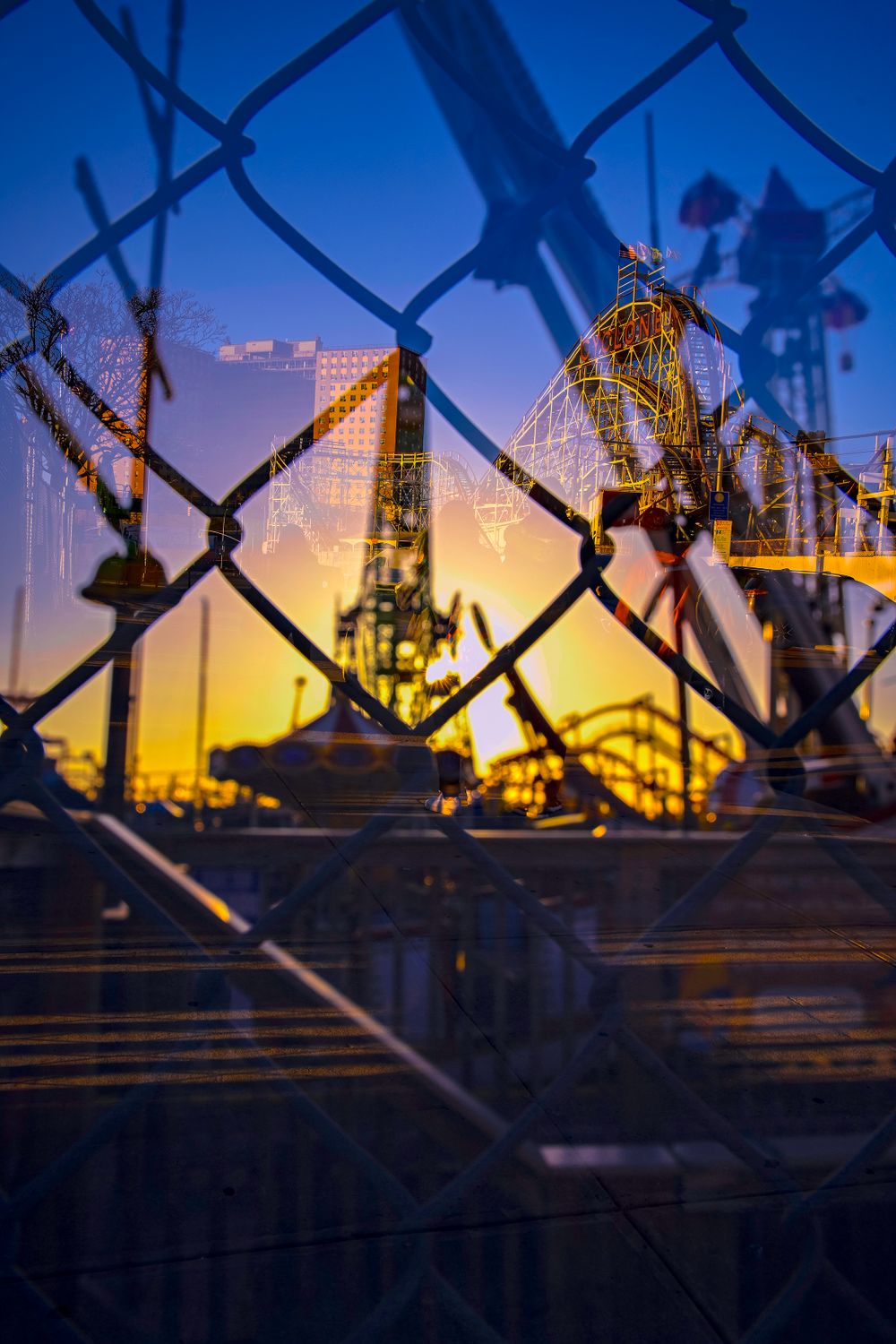 Chain-link fence frames sunset view of carnival rides at dusk.