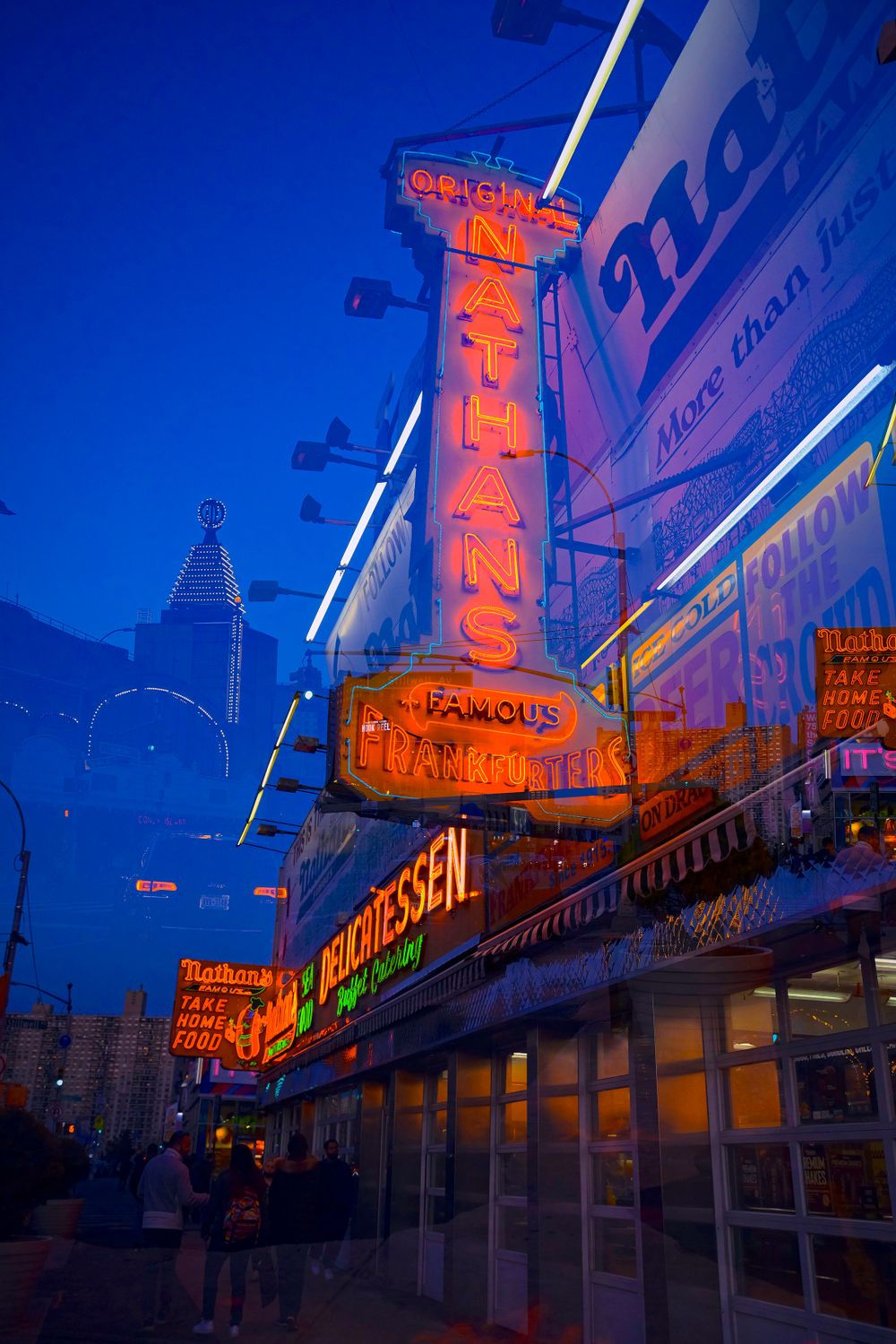 Neon signs illuminate the Nathan's Famous storefront at dusk in Coney Island.