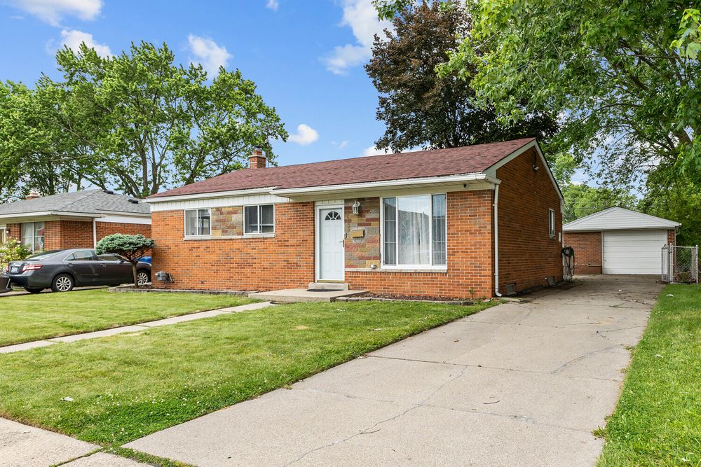 Brick ranch-style home with white entry door, attached driveway, detached garage, and green lawn.