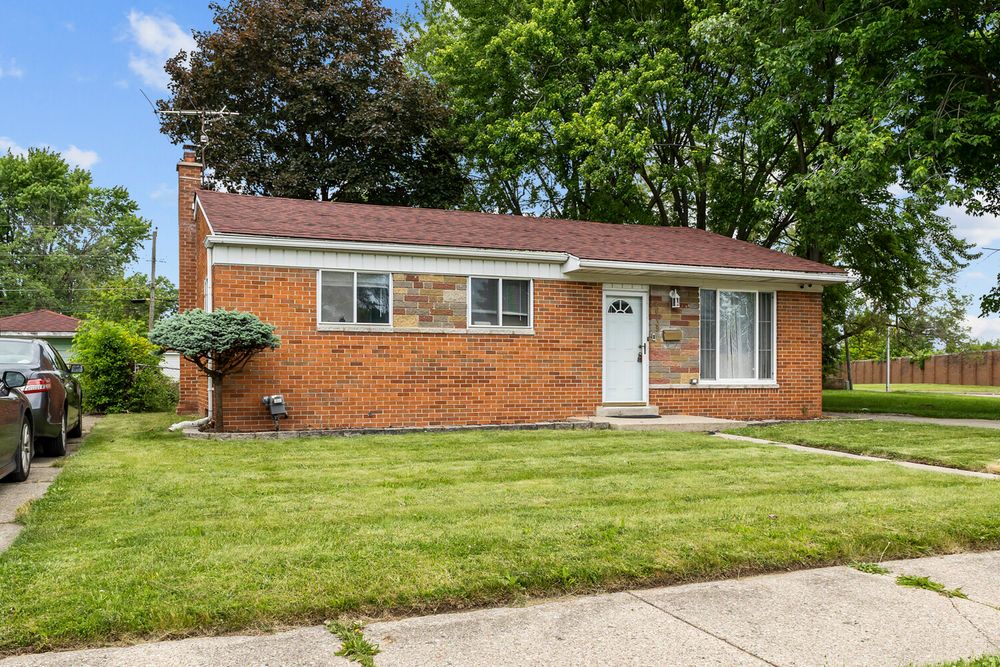 Brick ranch-style house with white entry door, well-maintained green lawn, and mature shade trees.
