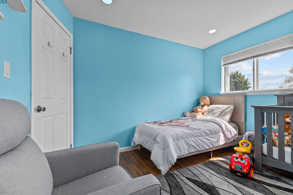 Blue childrens bedroom with bed, gray crib, gray armchair, and bright window.