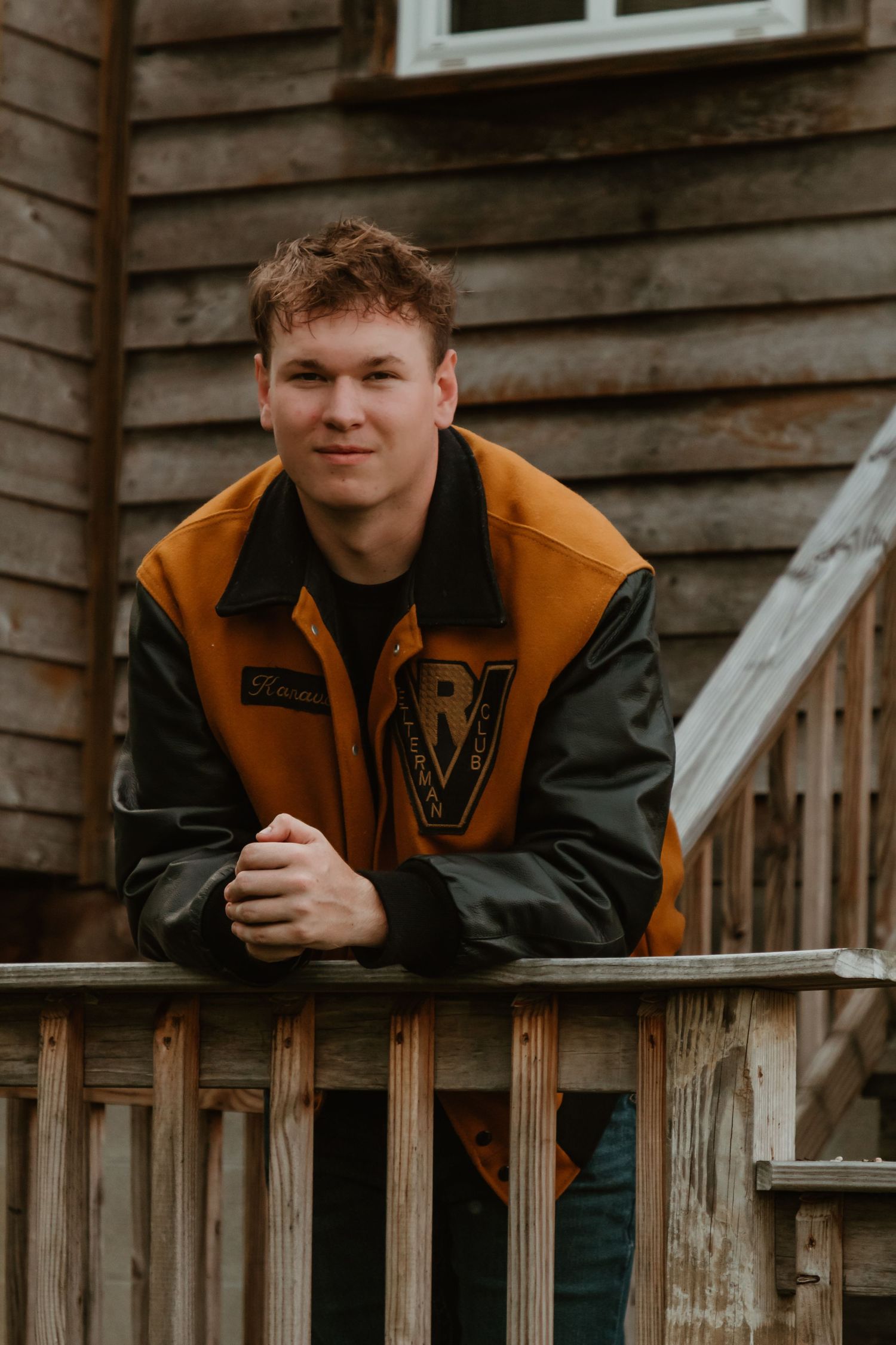 A person wearing a varsity-style jacket with orange and black colors leans on a wooden deck railing.