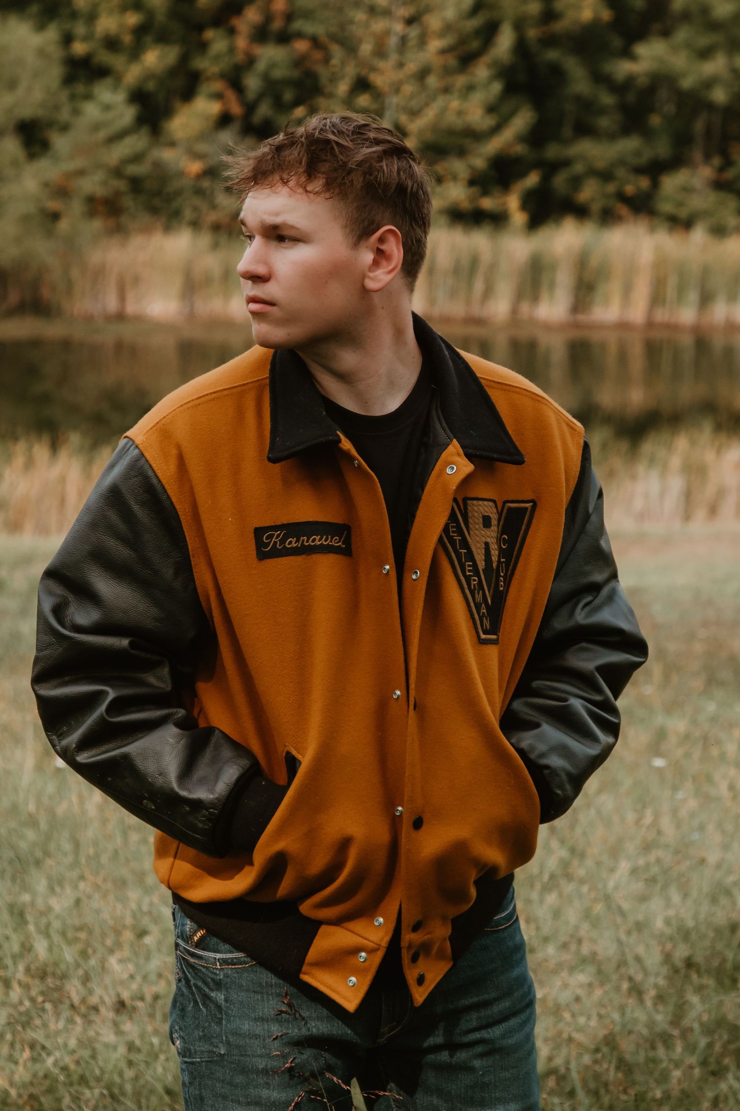 A person wearing a brown and black varsity letterman jacket poses outdoors in a natural setting.