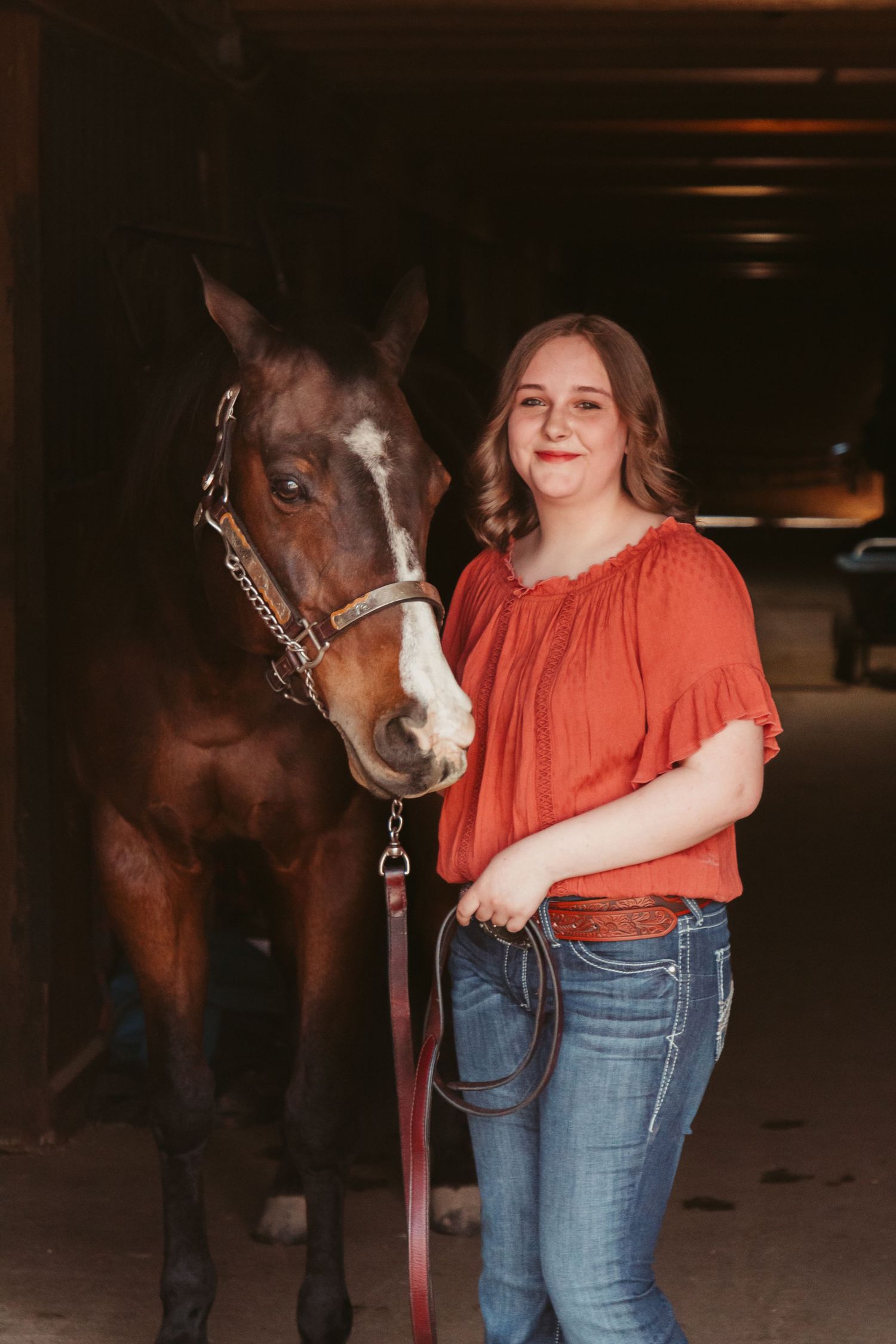 A person in an orange top and jeans stands next to a brown horse with a white blaze in a dimly lit stable area.