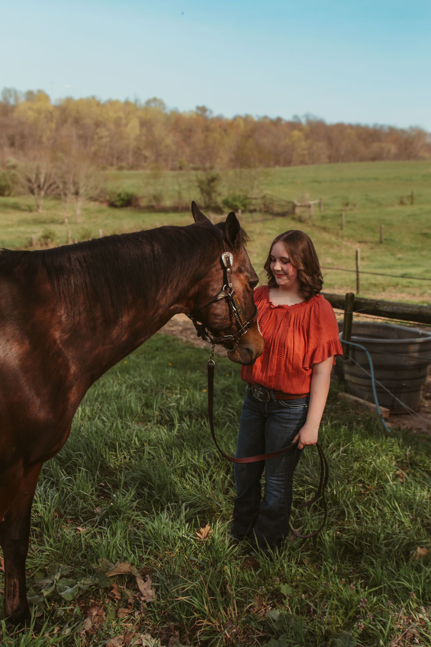 Woman in red blouse stands with dark brown horse in grassy field on autumn day.